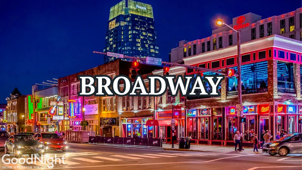 This energetic exterior photograph captures the iconic Broadway sign surrounded by the neon-lit facades of downtown Nashville. The lively atmosphere is emphasized by crowds of people, city traffic, and a backdrop of the city skyline, evoking the city’s busy nightlife and music scene.