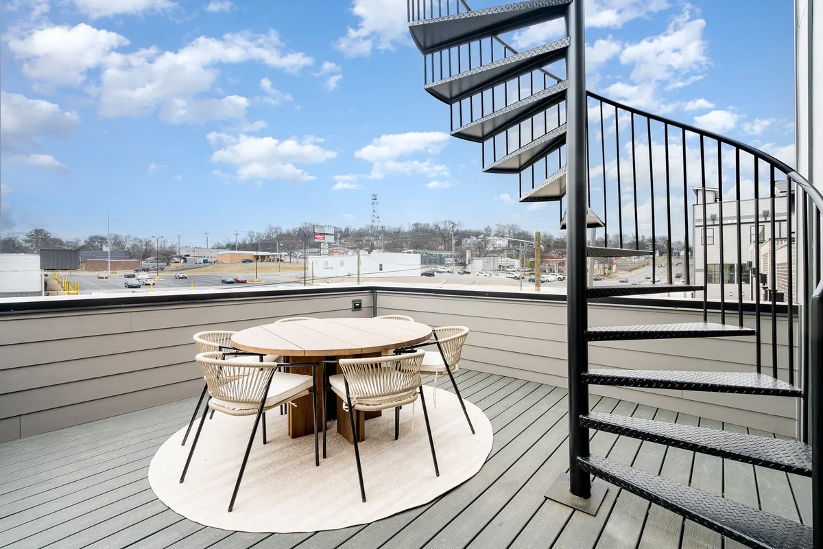 A beautiful rooftop patio featuring a spiral staircase connecting two levels. On the lower deck, there's a stylish outdoor dining table set for six and comfortable seating. The backdrop includes the Nashville skyline with warm sunset lighting, creating a relaxed and inviting atmosphere perfect for hosting meals or small gatherings.