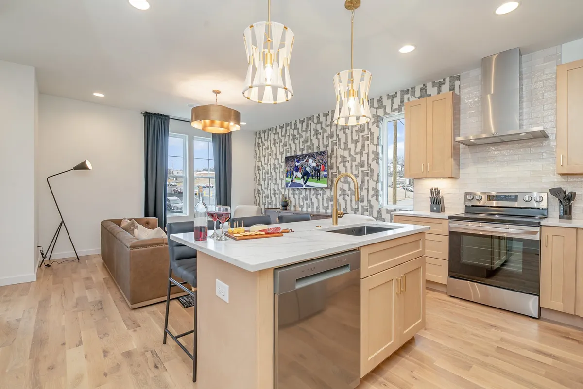 This image features a chic, open-concept living and kitchen area. A spacious kitchen island with bar seating anchors the room, while stylish hanging light fixtures illuminate the space. In the background, a plush sofa faces a wall-mounted Smart TV, and the dining area flows seamlessly into the living space. The overall ambiance is welcoming and contemporary, perfect for gatherings and group activities.