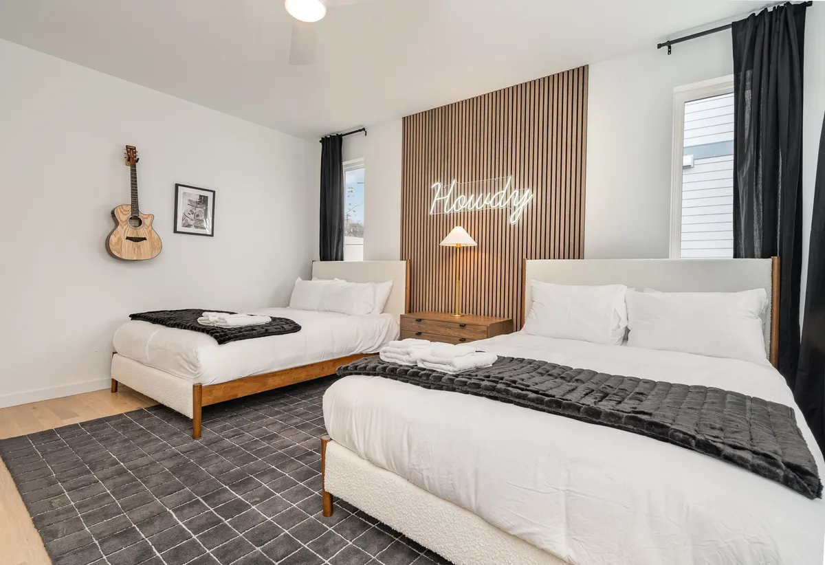 A contemporary bedroom featuring two beds with crisp white linens, a neon sign that adds personality, and a guitar mounted on the wall. The room is bright and inviting, perfect for families or friends sharing a space.