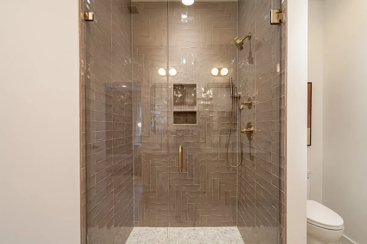 A modern bathroom with a glass shower enclosure, elegant herringbone tile, and gold fixtures, evoking a spa-like retreat. The design feels fresh, clean, and luxurious, perfect for relaxing after a day of travel.