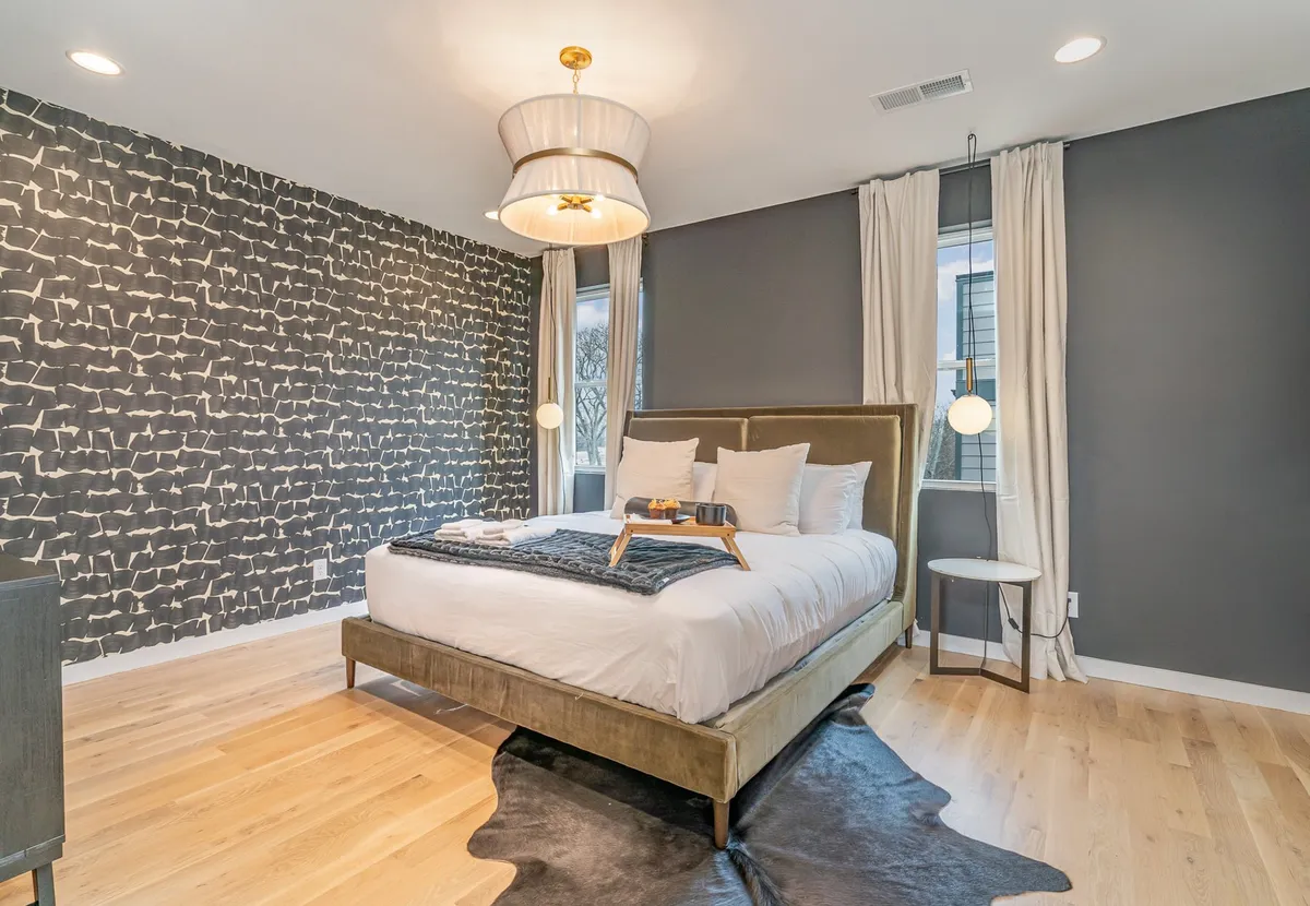 A serene, modern bedroom with a plush bed, textured decorative wall, stylish chandelier, and a side table with a lamp. Diffused natural light streams through unseen windows, creating a restful and inviting ambiance perfect for unwinding.