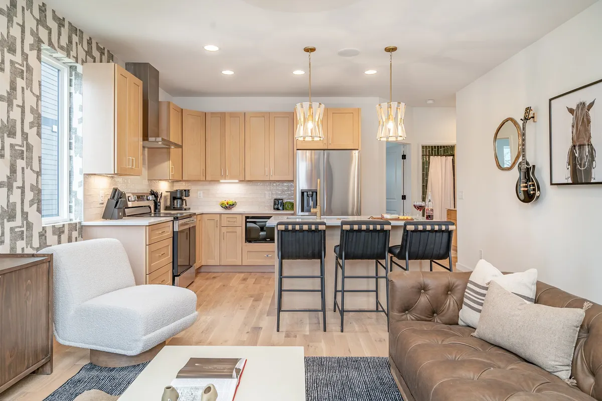 A warm and inviting interior shot capturing the communal hub of the home. The kitchen island with bar stools is a focal point, accompanied by a comfortable sofa in the foreground. The decorative wall pattern and modern light fixtures add flair, and the open layout connects cooking, relaxing, and entertaining areas.