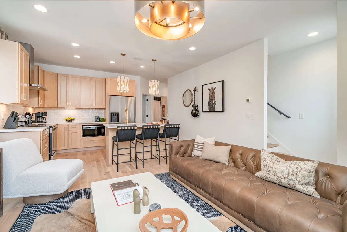 The image showcases a spacious, modern open-concept living area with a stylish sofa, inviting coffee table, sleek kitchen island, chic lighting fixtures, and curated wall art. The space is designed for socializing, whether guests are preparing meals together, hosting game nights, or unwinding after exploring Nashville.